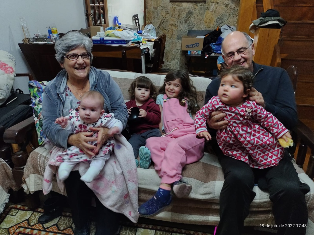Na imagem há um casal de idosos com quatro crianças. Duas crianças estão sentadas no sofá e dois bebês estão no colo dos adultos. Eles estão sorrindo. 